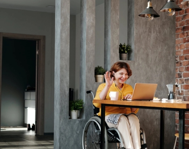 a lady in a wheelchair sitting at a table with a coffee. She's looking at a computer screen and waving to someone she's obviously having a video call with.