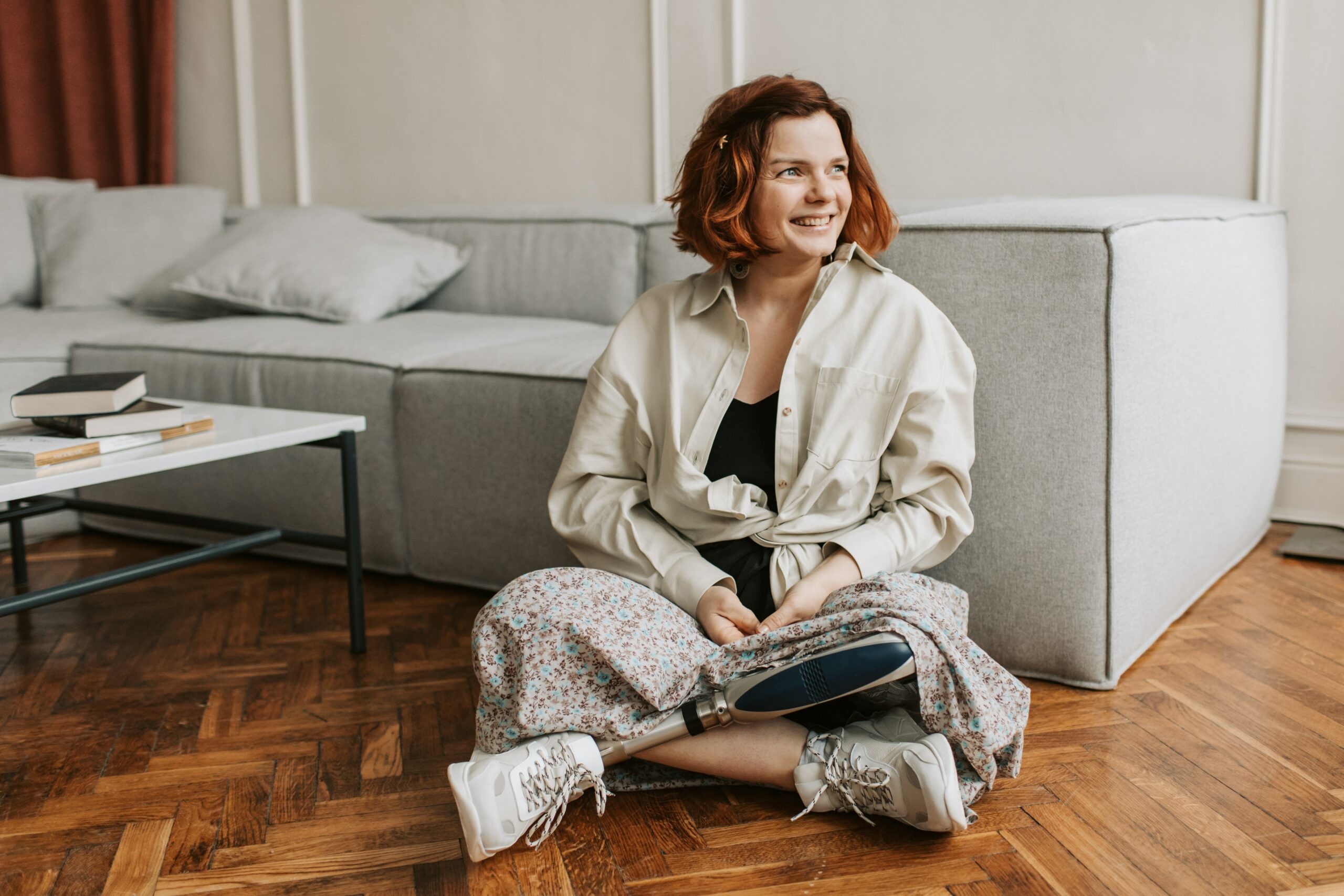 This is a photo a happy woman with a prosthetic leg sitting cross-legged on the floor of her home, against her sofa, smiling and looking out of the window, which is out of shot.